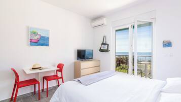Habitación de hotel con cama, escritorio con televisor, una silla roja, pared blanca con un cuadro y balcón con vistas.