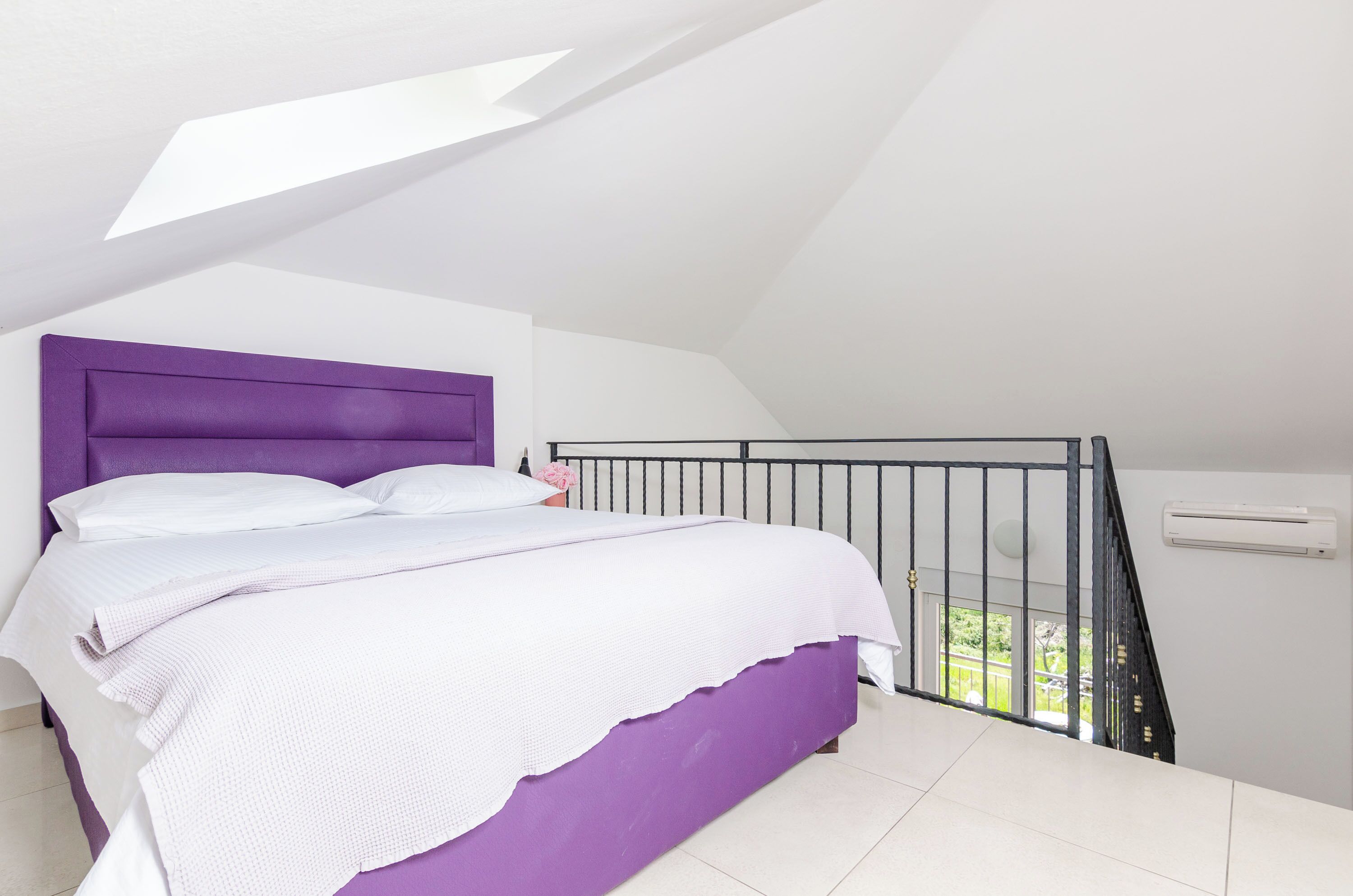 A bedroom with a purple headboard, white bedding, and a view of the outdoors through a window.