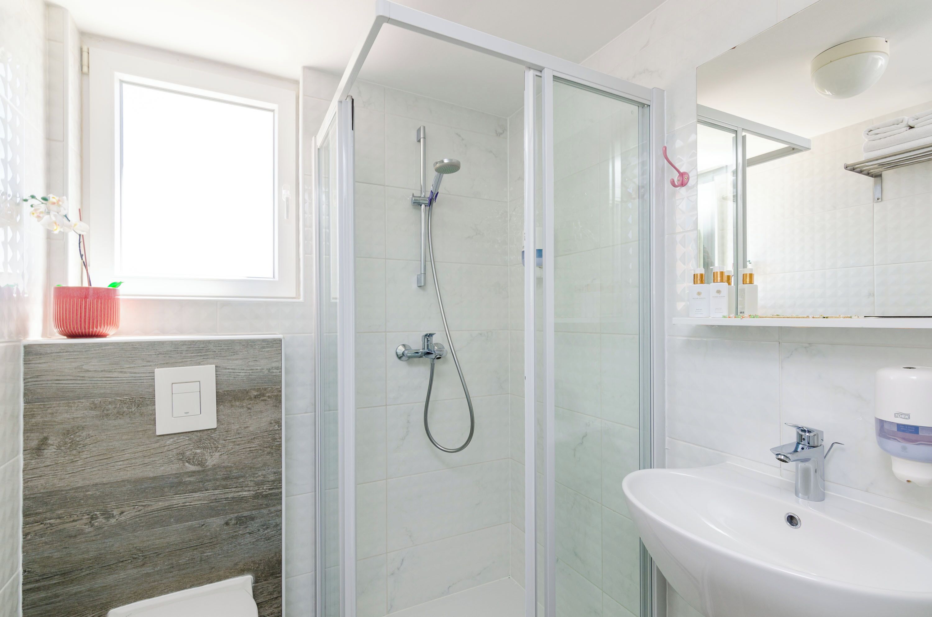 Un baño moderno con ducha envuelta en vidrio, un lavabo blanco y una ventana con una planta.