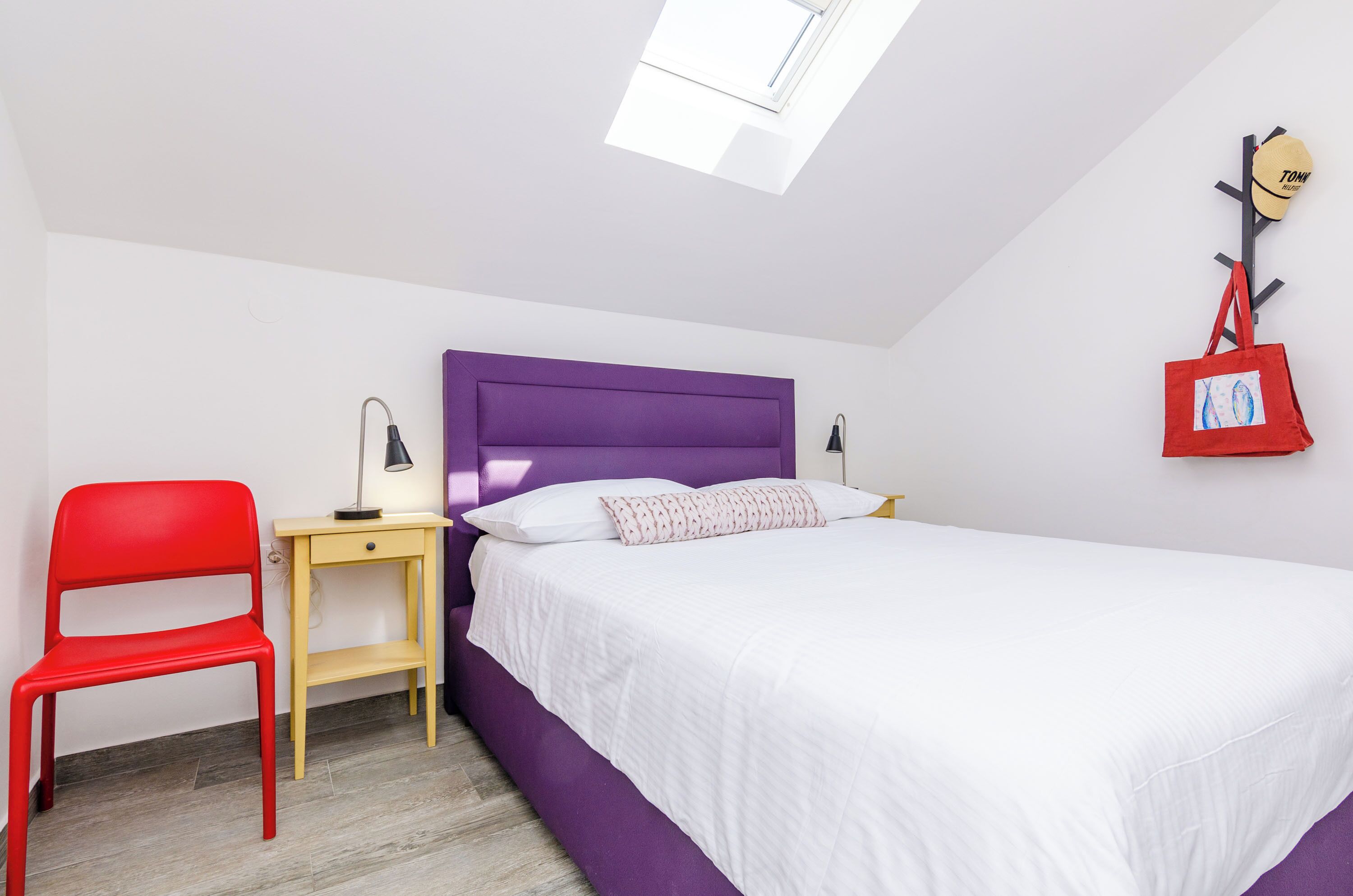 A bedroom with a purple headboard, a white bed, a red chair, a small table with a lamp, and a red bag hanging on the wall.