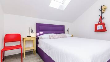 A bedroom with a purple headboard, a white bed, a red chair, a small table with a lamp, and a red bag hanging on the wall.