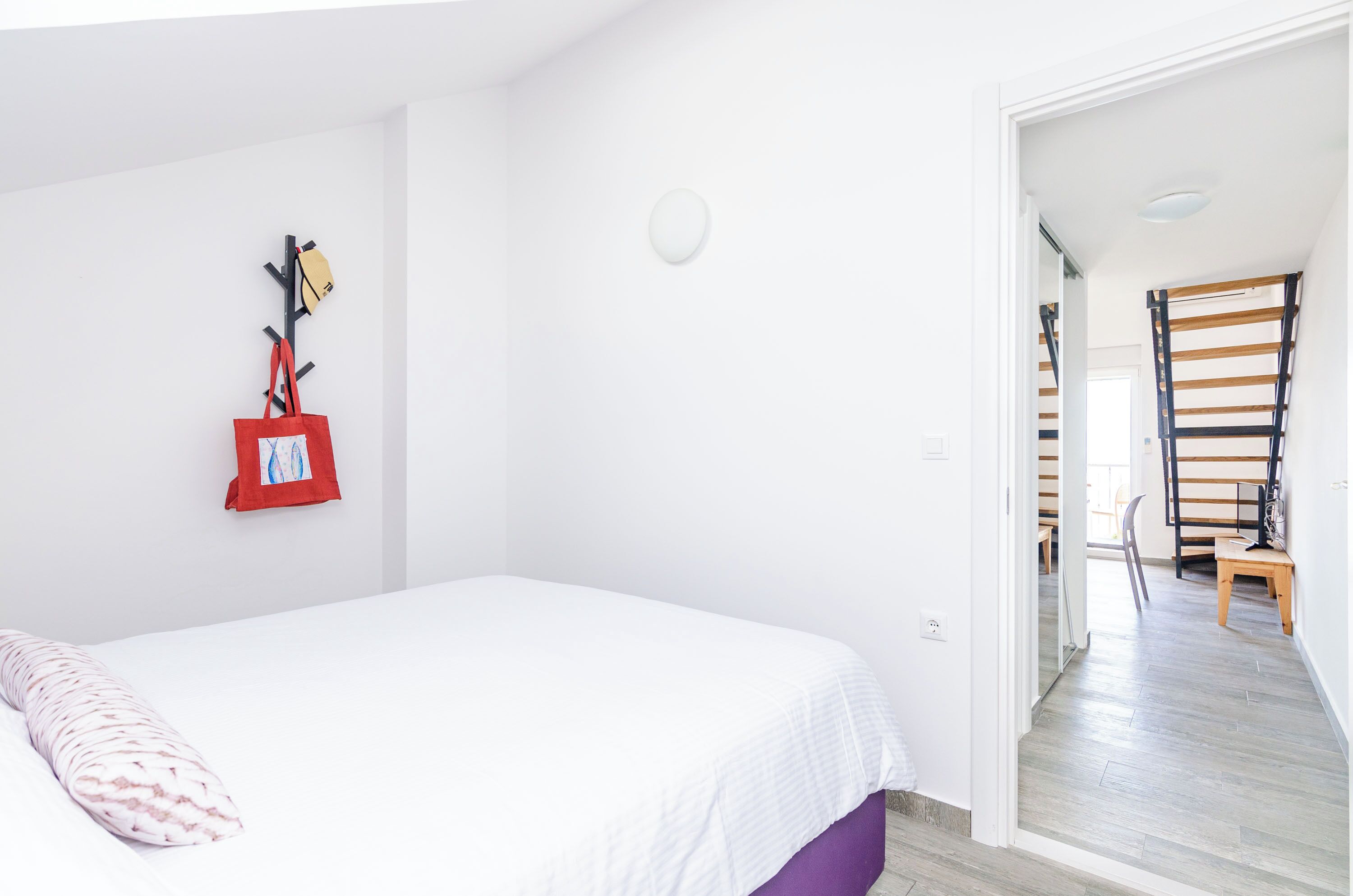 A bedroom with a white bed, a red bag hanging on a wall-mounted rack, and a wooden chair in the adjacent room.