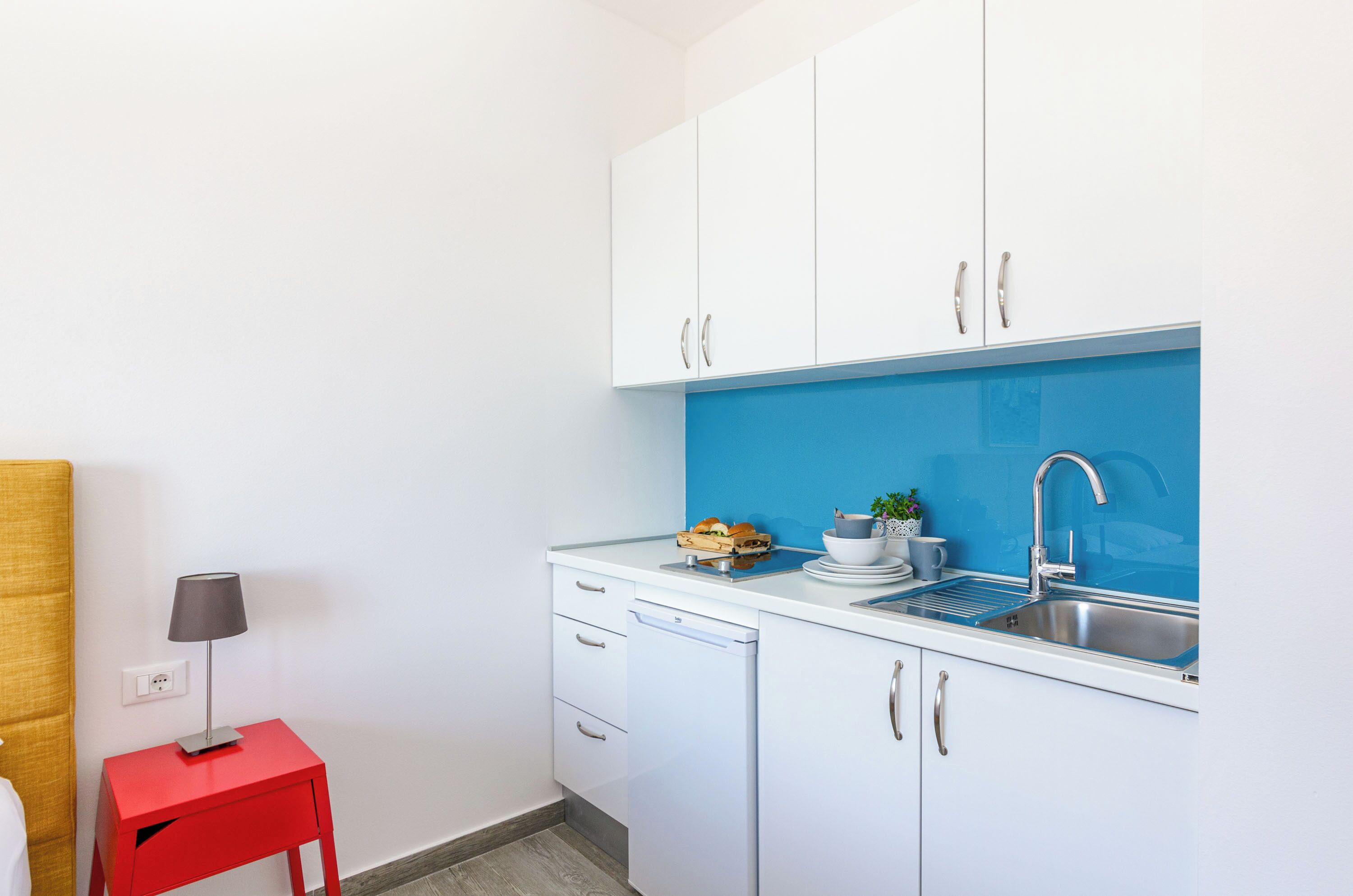 Cocina compacta con armarios blancos, salpicadero azul y una mesita roja con una lámpara.