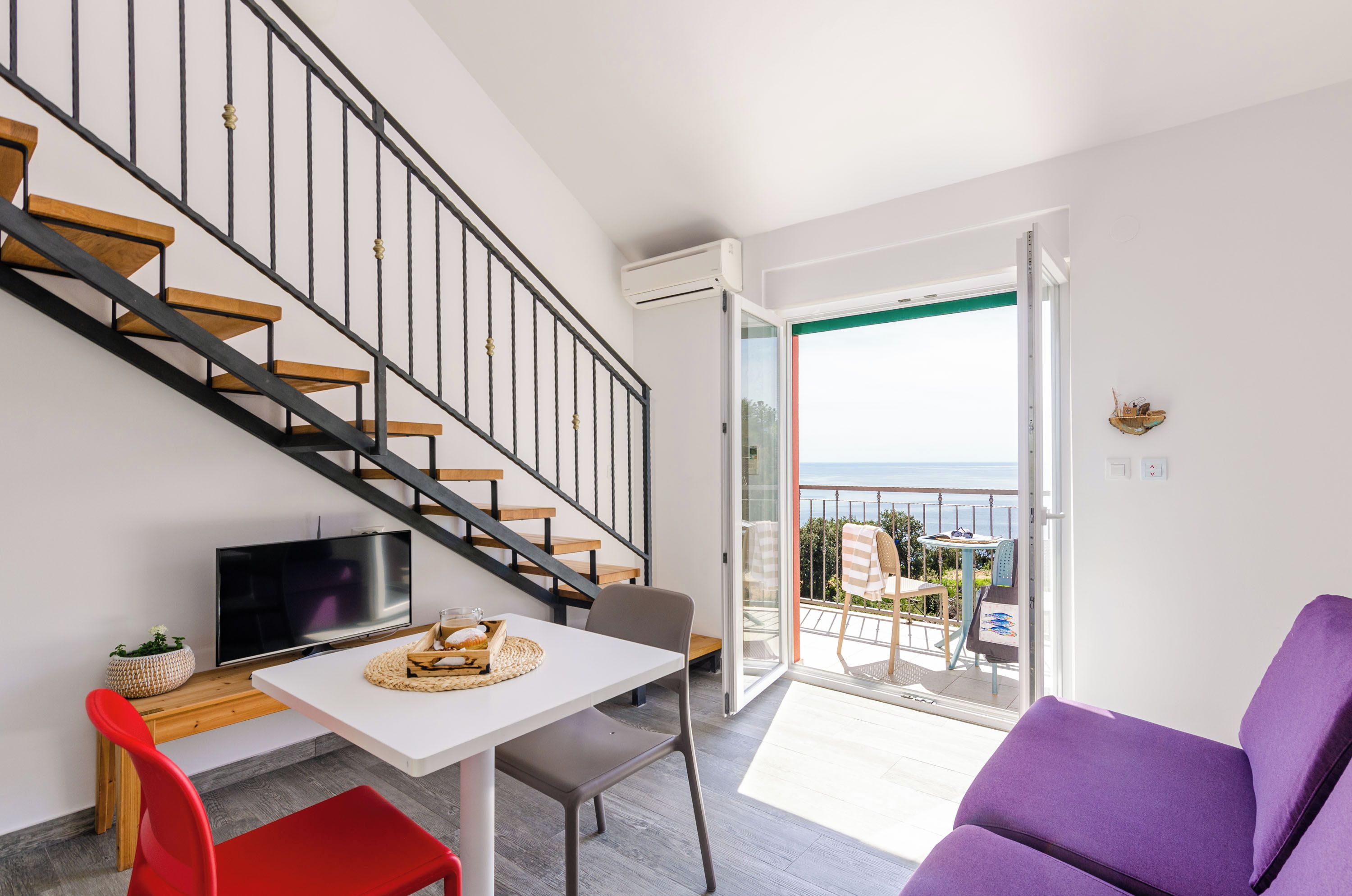 A modern apartment with a staircase, a dining area, a living room with a purple sofa, and a balcony with a view.