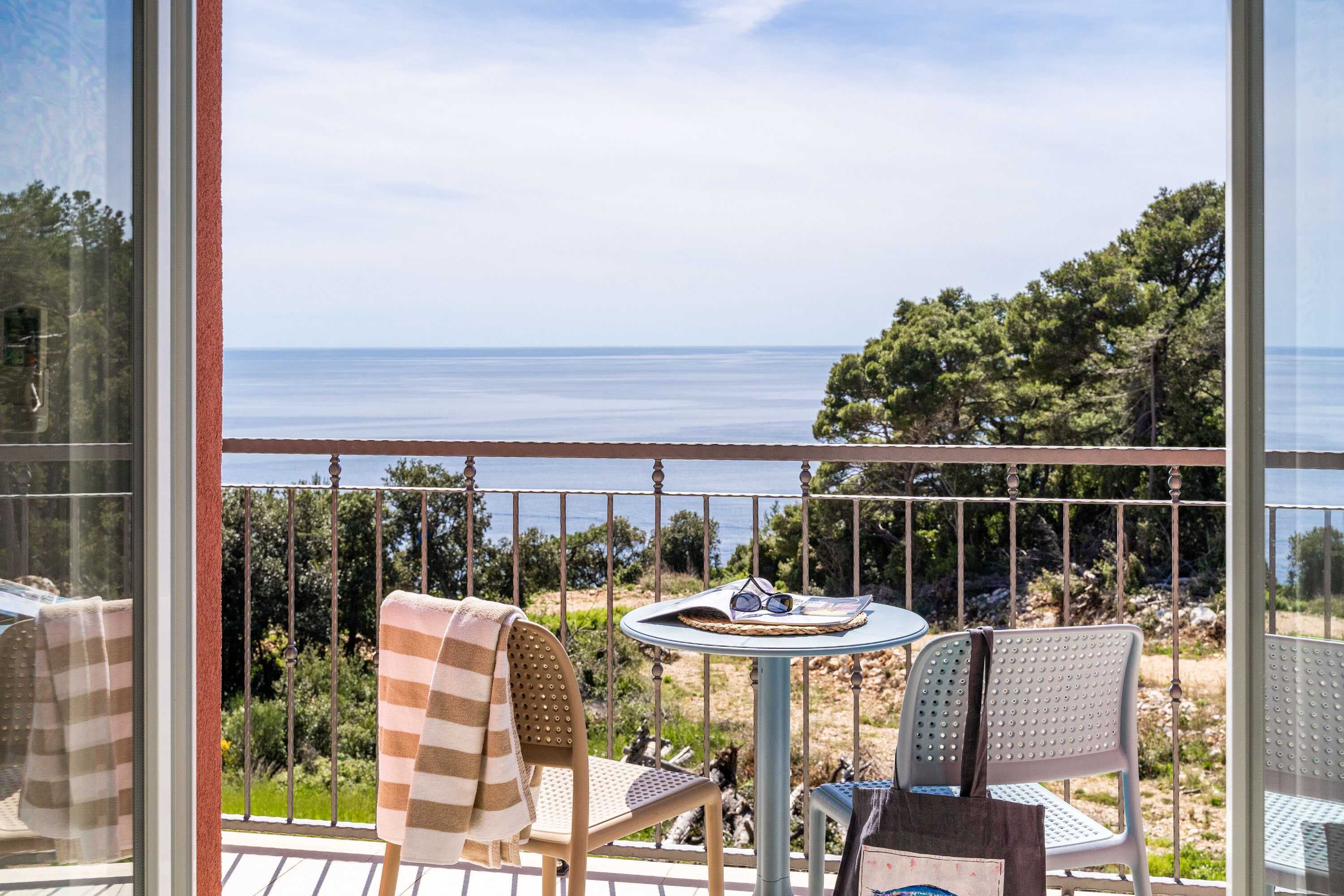 A balcony with a table and chairs, overlooking a coastal landscape with trees and the sea.