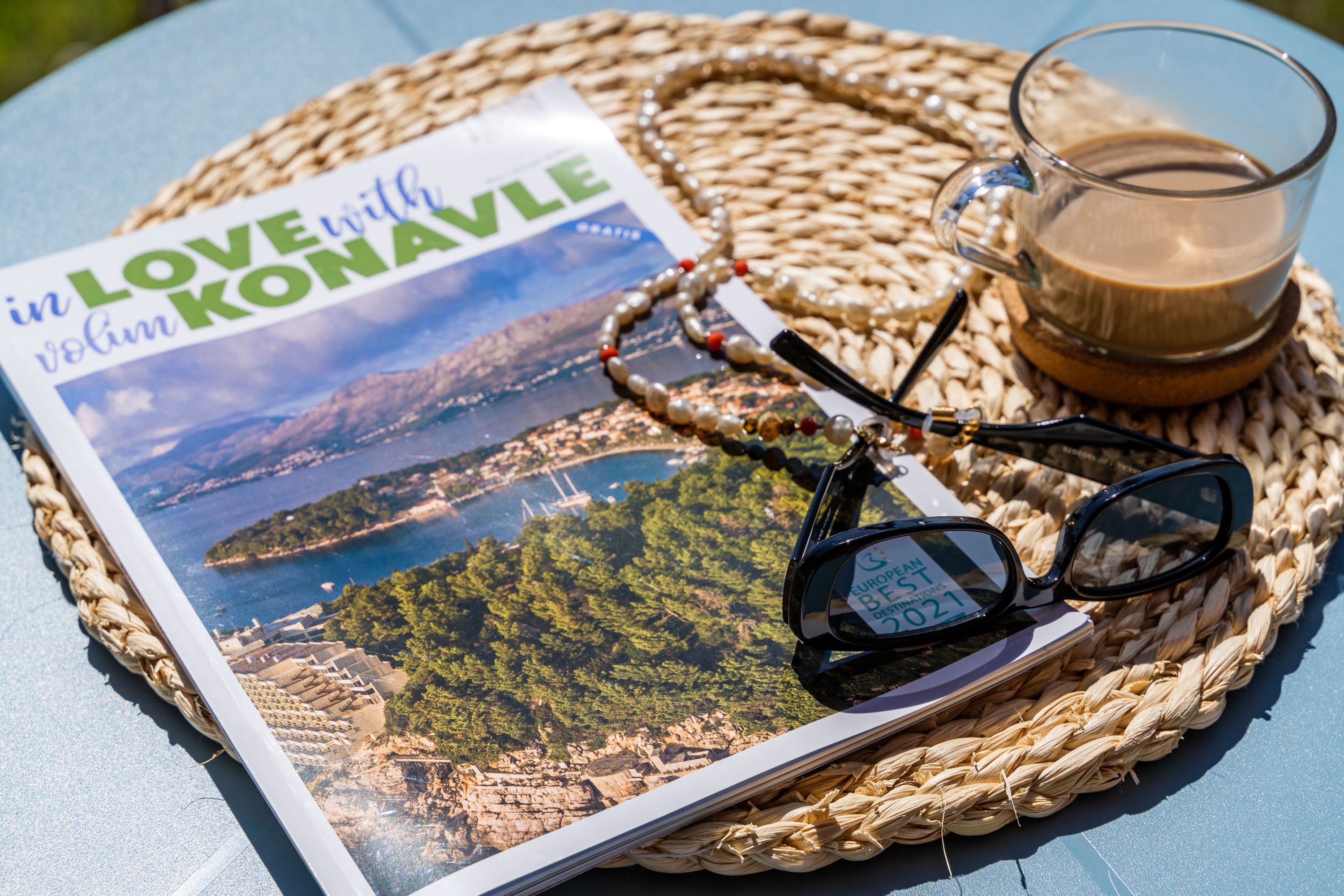Una revista titulada 'in LOVE with Konavle', un par de gafas y una taza de café sobre un tapete tejido.