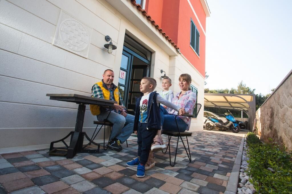 A family of four seated at an outdoor table with a tiled patio, a parked scooter, and a building with a red facade and a blue door.