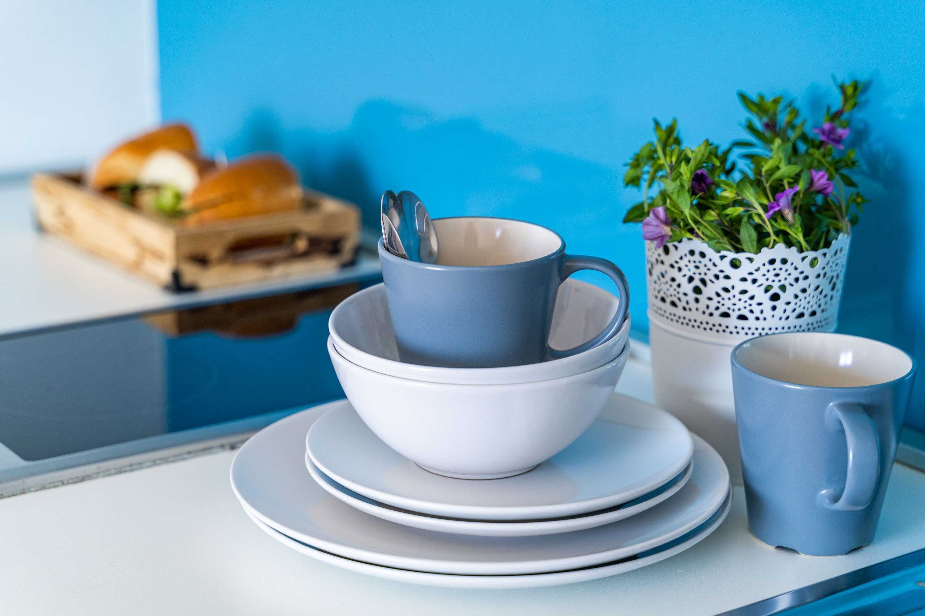 Une table avec une pile d’assiettes blanches, une tasse bleue et un bol blanc avec une cuillère. En arrière-plan, on aperçoit une caisse en bois contenant un sandwich et un pot blanc avec des fleurs violettes.