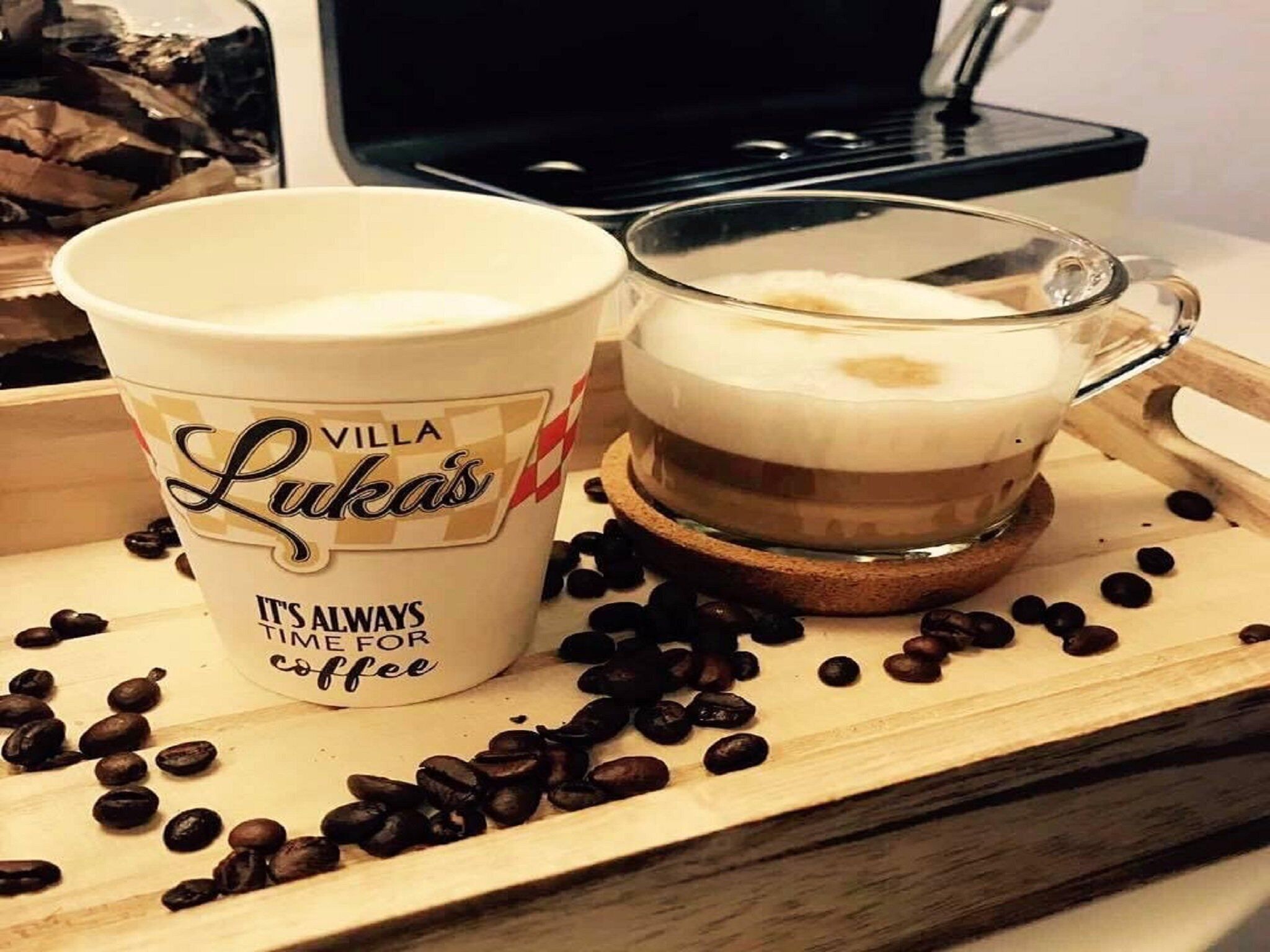 A cup of coffee with a branded mug and a glass cup on a wooden tray with coffee beans.