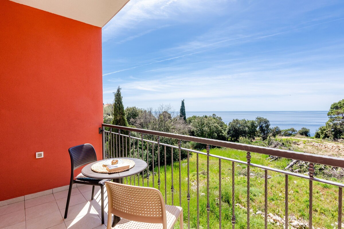 A balcony with a table and chairs, overlooking a coastal landscape with trees and the sea.