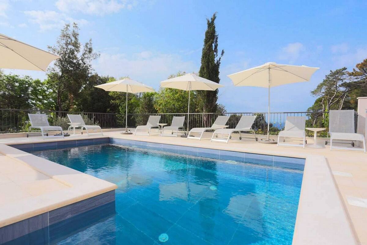 A swimming pool with lounge chairs and umbrellas, surrounded by trees and a clear sky.