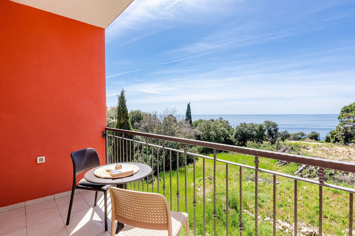 A balcony with a table and chairs, overlooking a coastal landscape with trees and the sea.