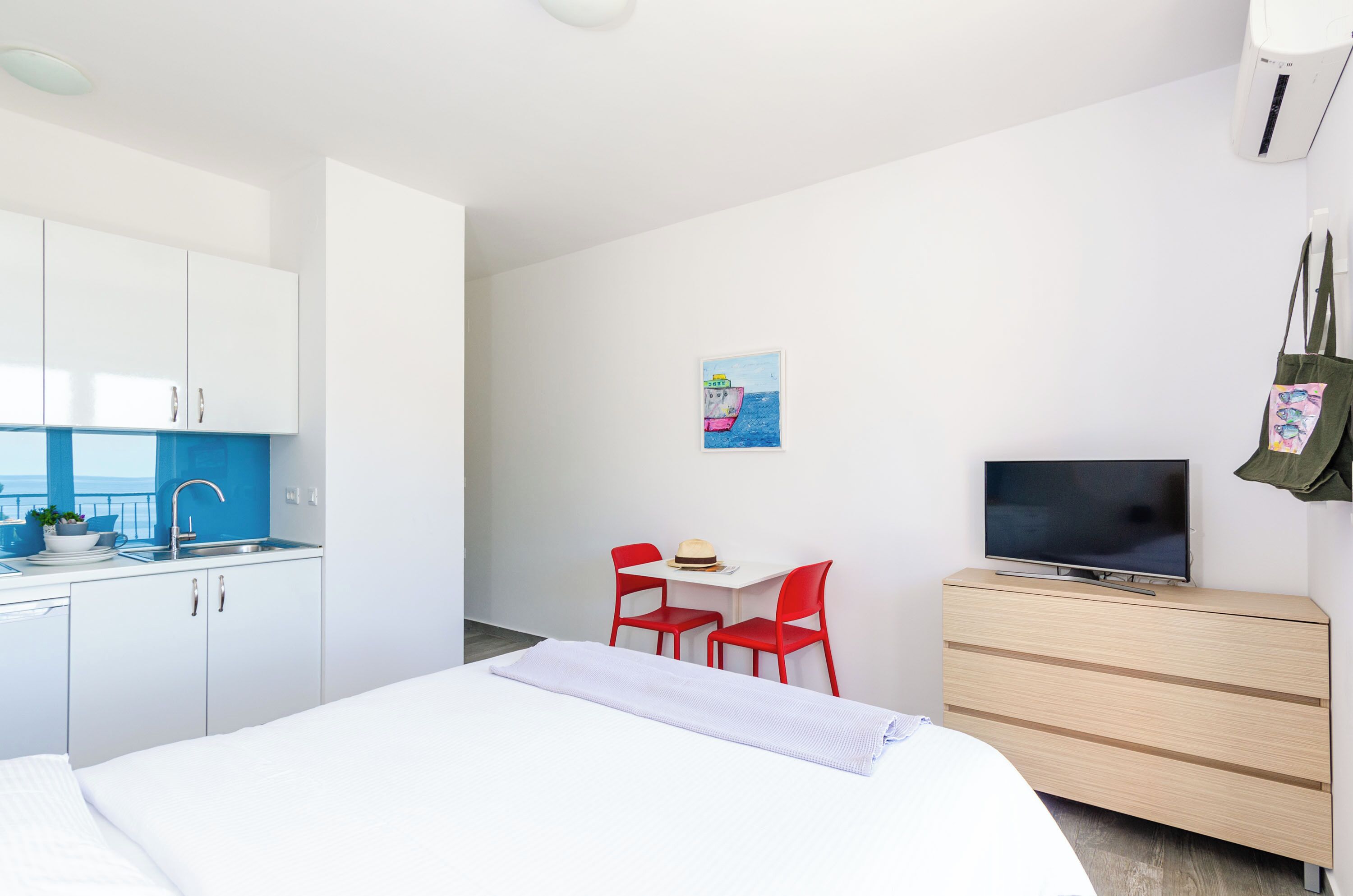 A modern hotel room with a bed, a desk with two red chairs, a TV on a wooden dresser, and a small painting on the wall.