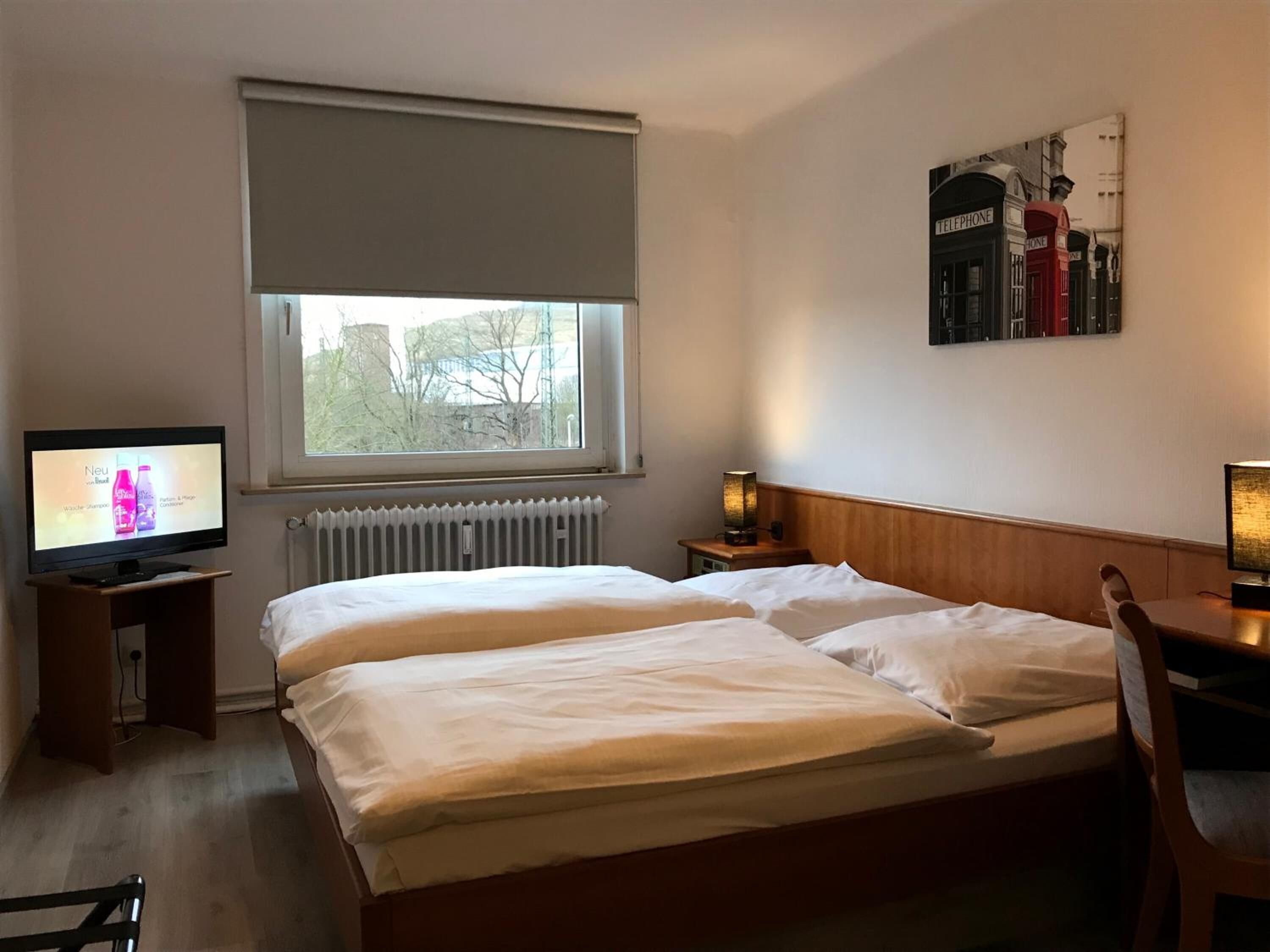 A hotel room with a bed, a television, a radiator, and a window with a view of trees.