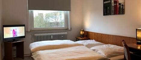 A hotel room with a bed, a television, a radiator, and a window with a view of trees.