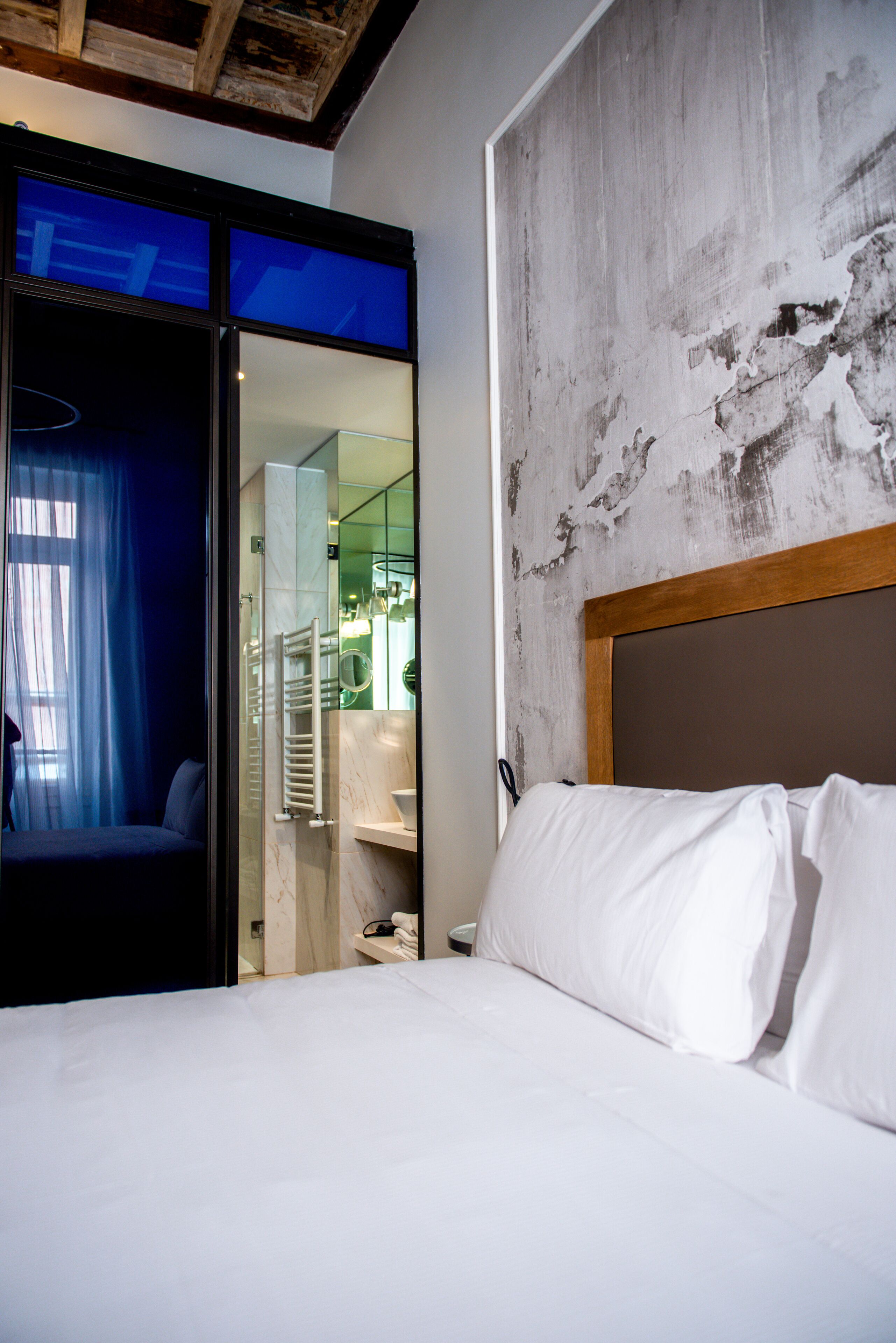 Une chambre d’hôtel avec un lit, une salle de bain visible à travers une porte en verre, et un design moderne avec un mur en béton et une tête de lit en bois.
