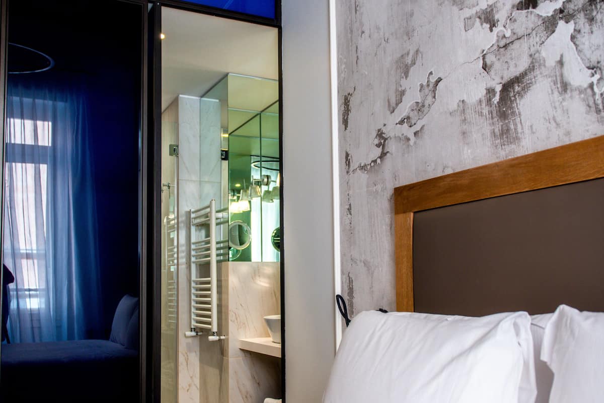 A hotel room with a bed, a bathroom visible through a glass door, and a modern design with a concrete wall and wooden headboard.