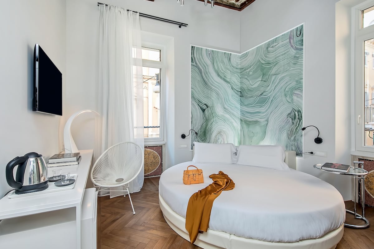 Ein modernes Schlafzimmer mit einem großen Bett, einem Flachbildfernseher, einem Schreibtisch mit Wasserkocher, einem Sessel und einem großen abstrakten Gemälde an der Wand.