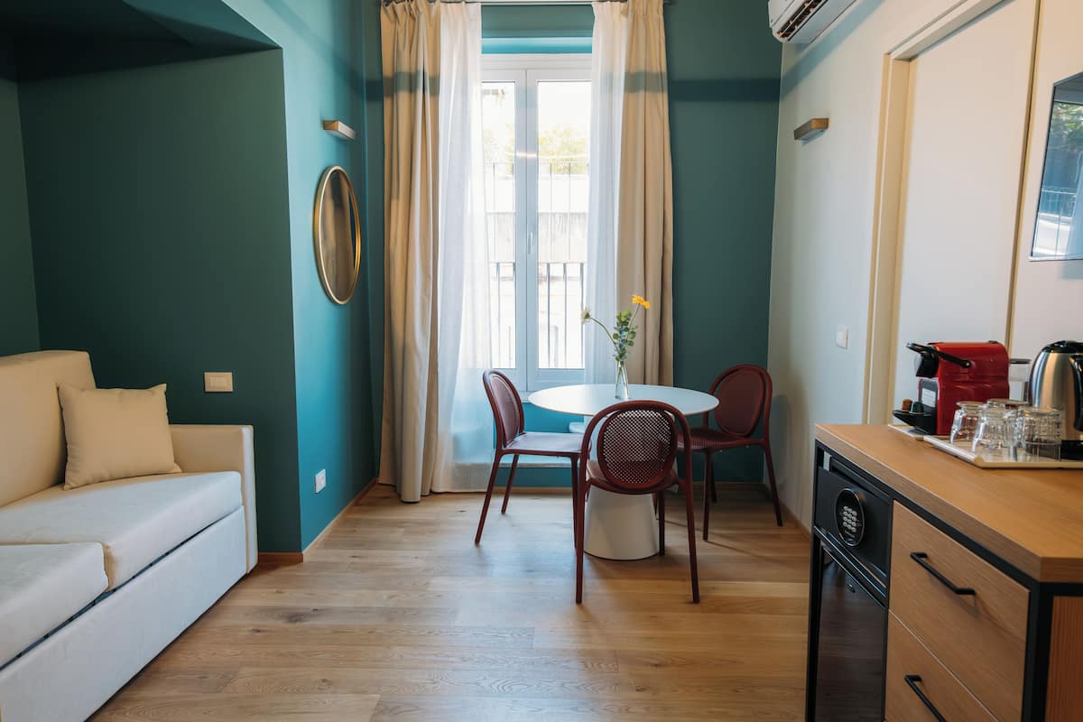 Ein Zimmer mit türkisfarbenen Wänden, Holzboden, einem weißen Sofa, einem Esstisch mit Stühlen und einer Küchenzeile inklusive Kaffeemaschine.