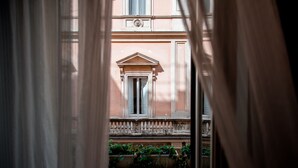 Une vue à travers des rideaux légers d’un bâtiment doté d’un balcon, d’une fenêtre avec des volets et d’une balustrade décorative.
