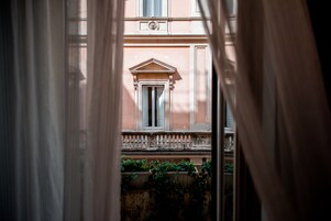 Blick durch transparente Vorhänge auf ein Gebäude mit Balkon, einem Fenster mit Rollläden und einem dekorativen Geländer.
