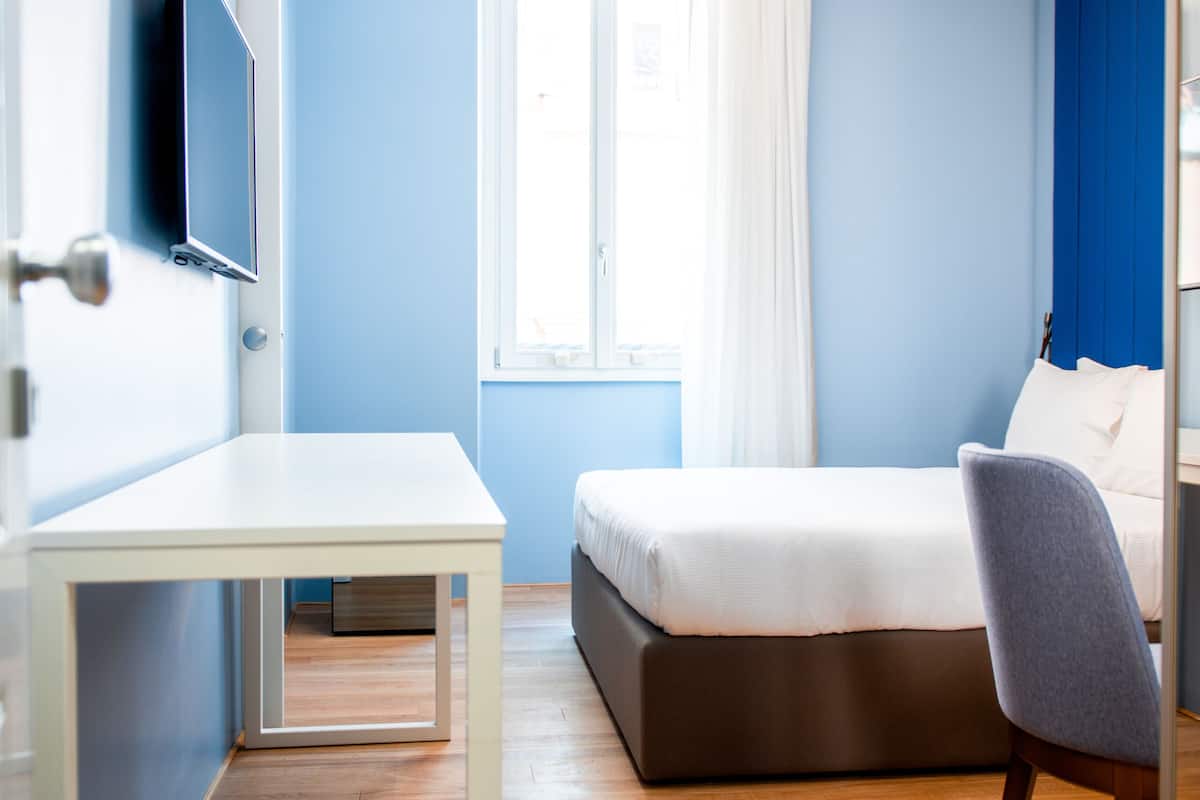Ein Zimmer mit einem Bett, einem Schreibtisch, einem Sessel, einem Fernseher und einem fenster mit Vorhängen.