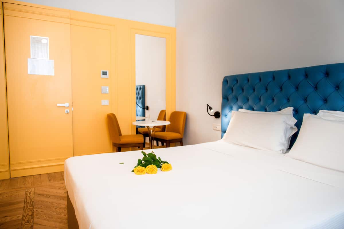 Ein Hotelzimmer mit einem Bett, einer blauen Kopfteil, einem kleinen Tisch mit Stühlen und einer Tür mit Spiegel und Lichtschalter.