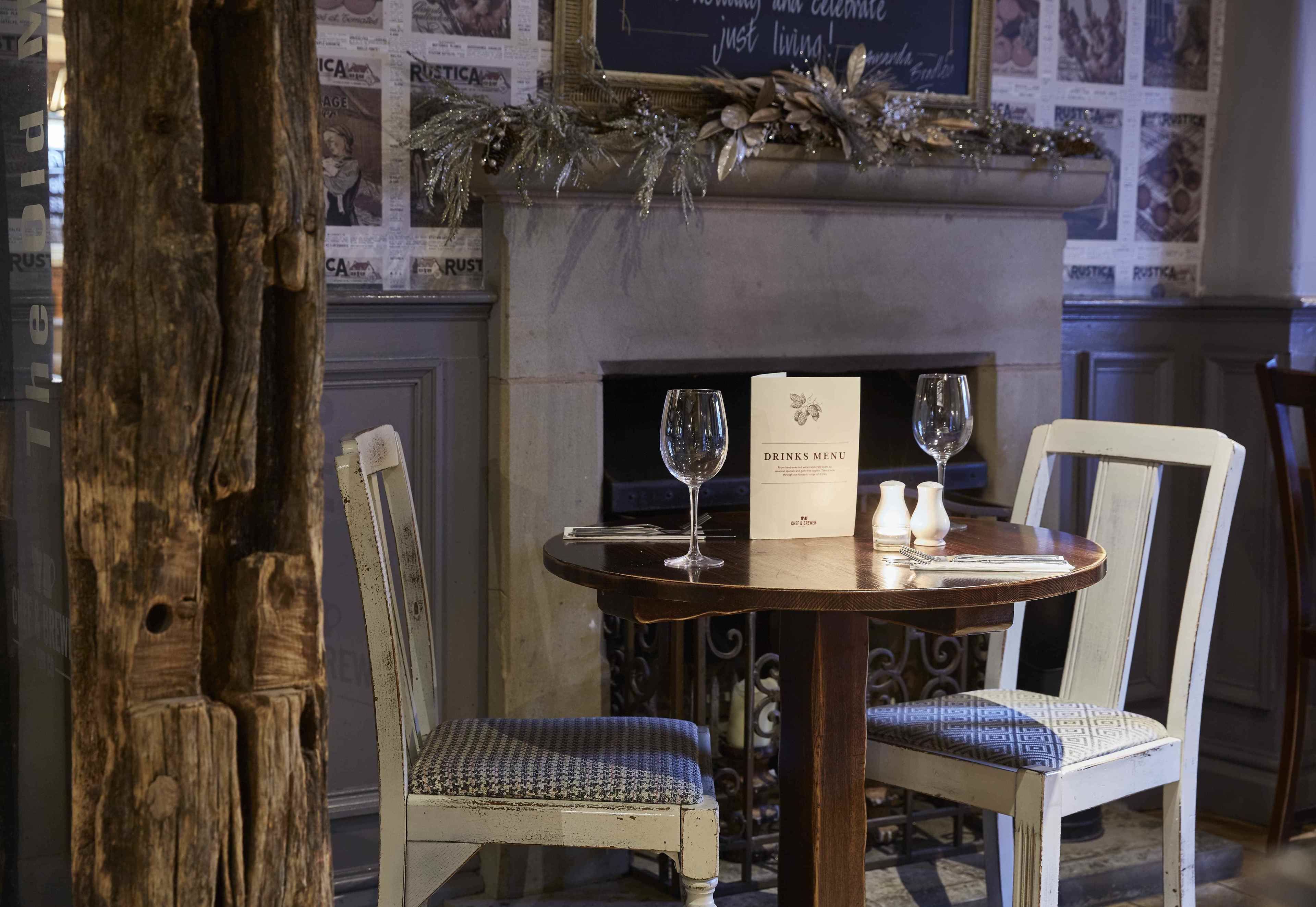 A dining table with two wine glasses and a menu, set against a fireplace with decorative garlands.