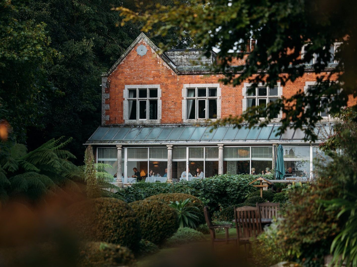 A brick building with a large glass front, surrounded by lush greenery.