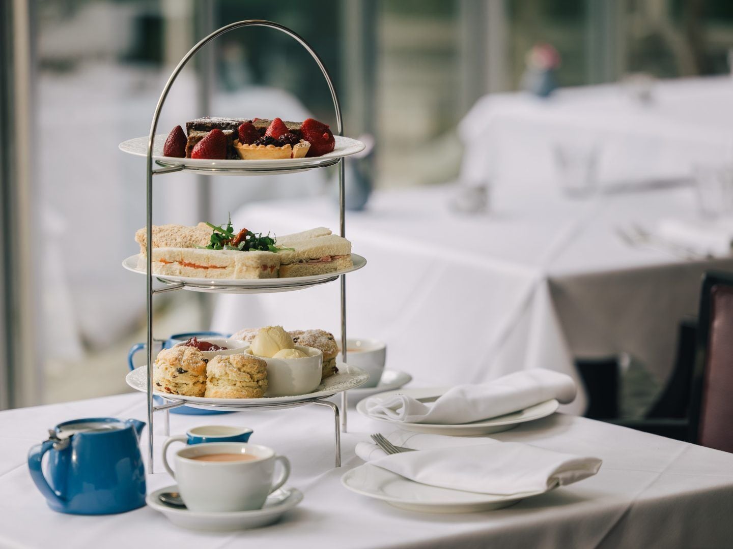 Ein dreistöckiges Kuchenregal mit Sandwiches, Scones und Gebäck auf einem Tisch mit Teetassen und Kaffeetassen.