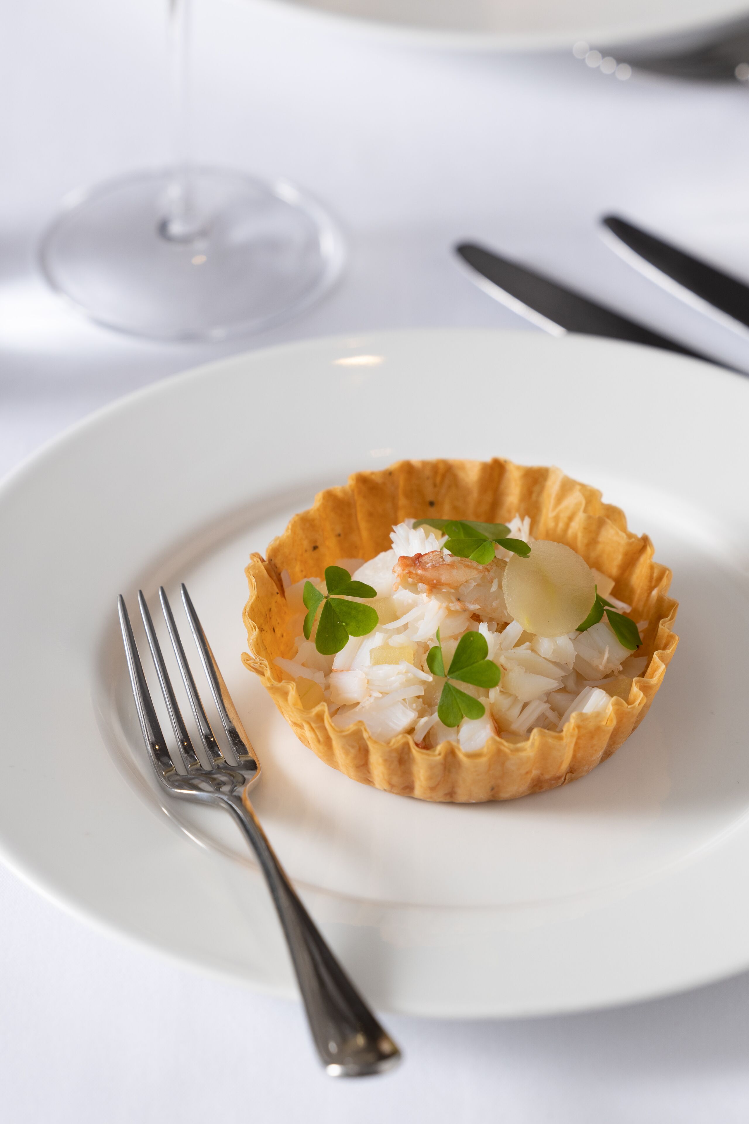A white plate with a seafood dish in a tart shell, accompanied by a fork and a glass of white wine.