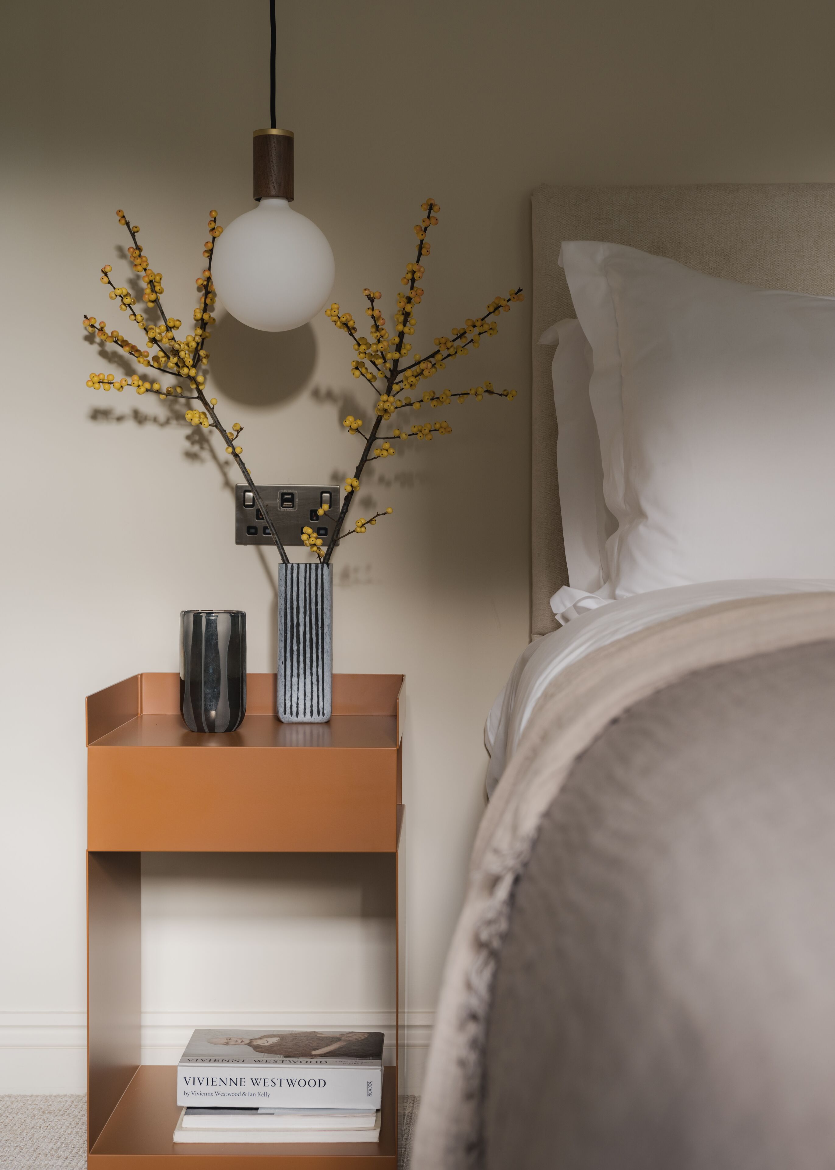 A neatly arranged bedside table with a lamp, a vase with yellow flowers, and a book titled "Vivienne Westwood".