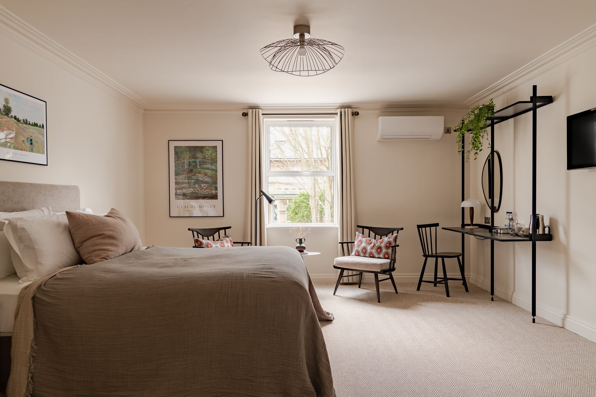A bedroom with a large bed, a desk, two chairs, a hanging plant, and a window with curtains.