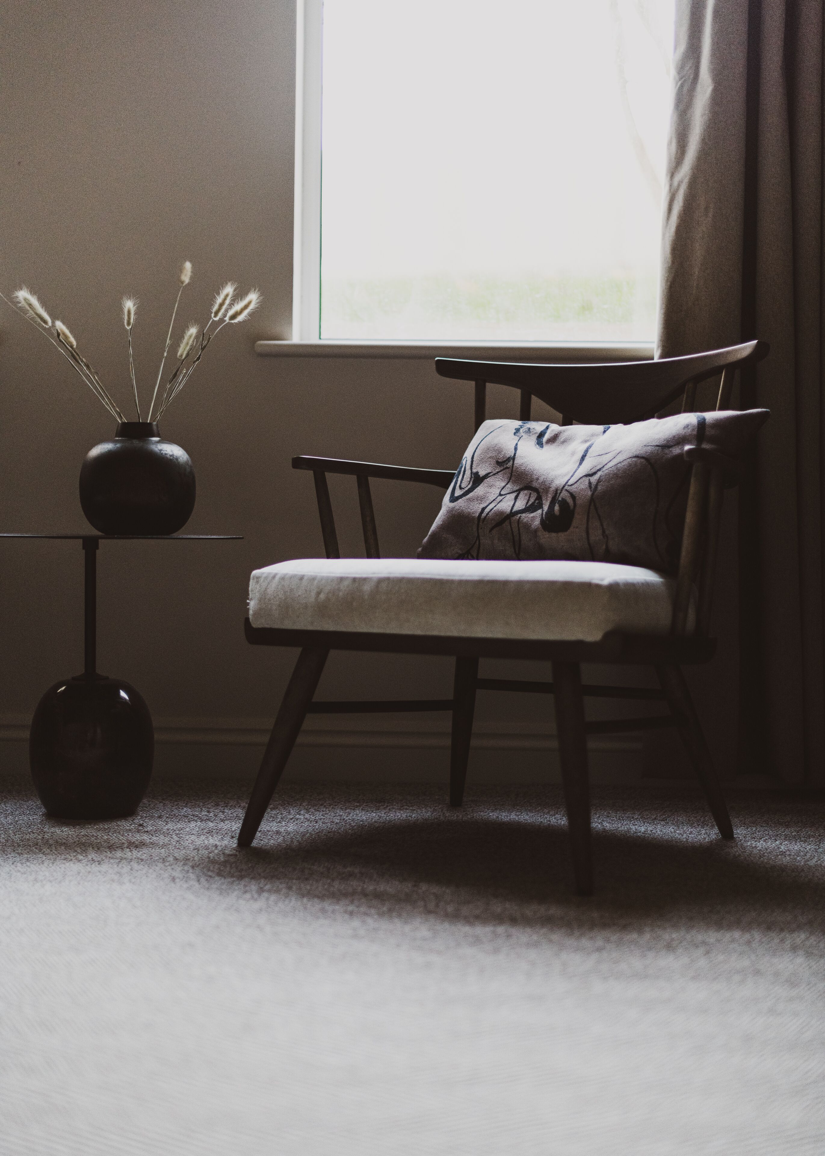 Une chaise avec un coussin et un oreiller, un vase contenant des plantes séchées et une petite table sur laquelle se trouve un objet sphérique.