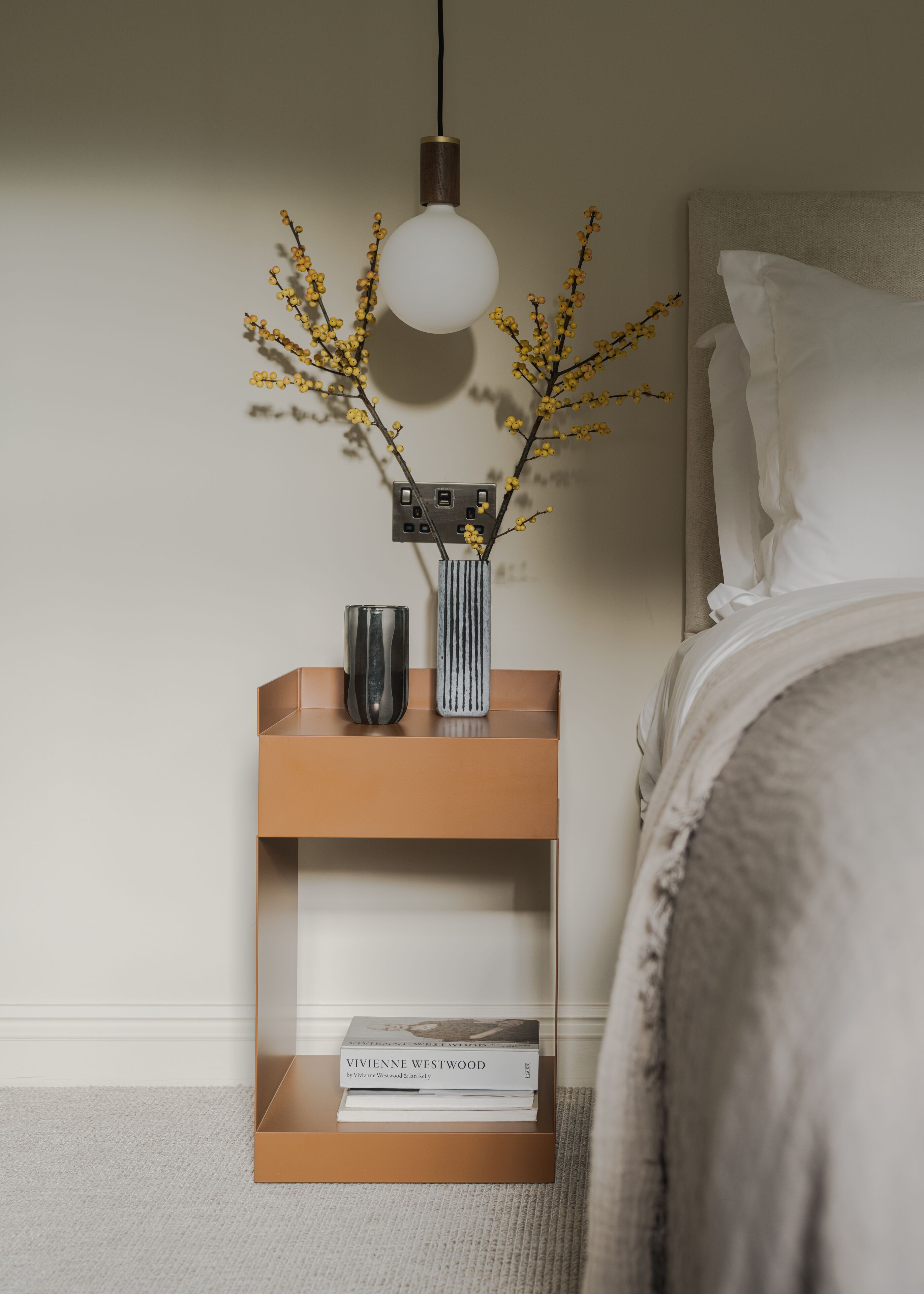 A neatly arranged bedside table with a lamp, a vase with yellow flowers, and a book titled "Vivienne Westwood" by Vivienne Westwood & Im Kelly.