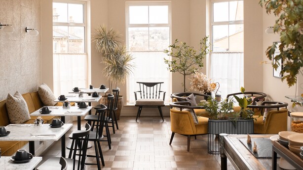 Uma sala de jantar ampla, com várias mesas e cadeiras, janelas grandes e plantas em vasos.
