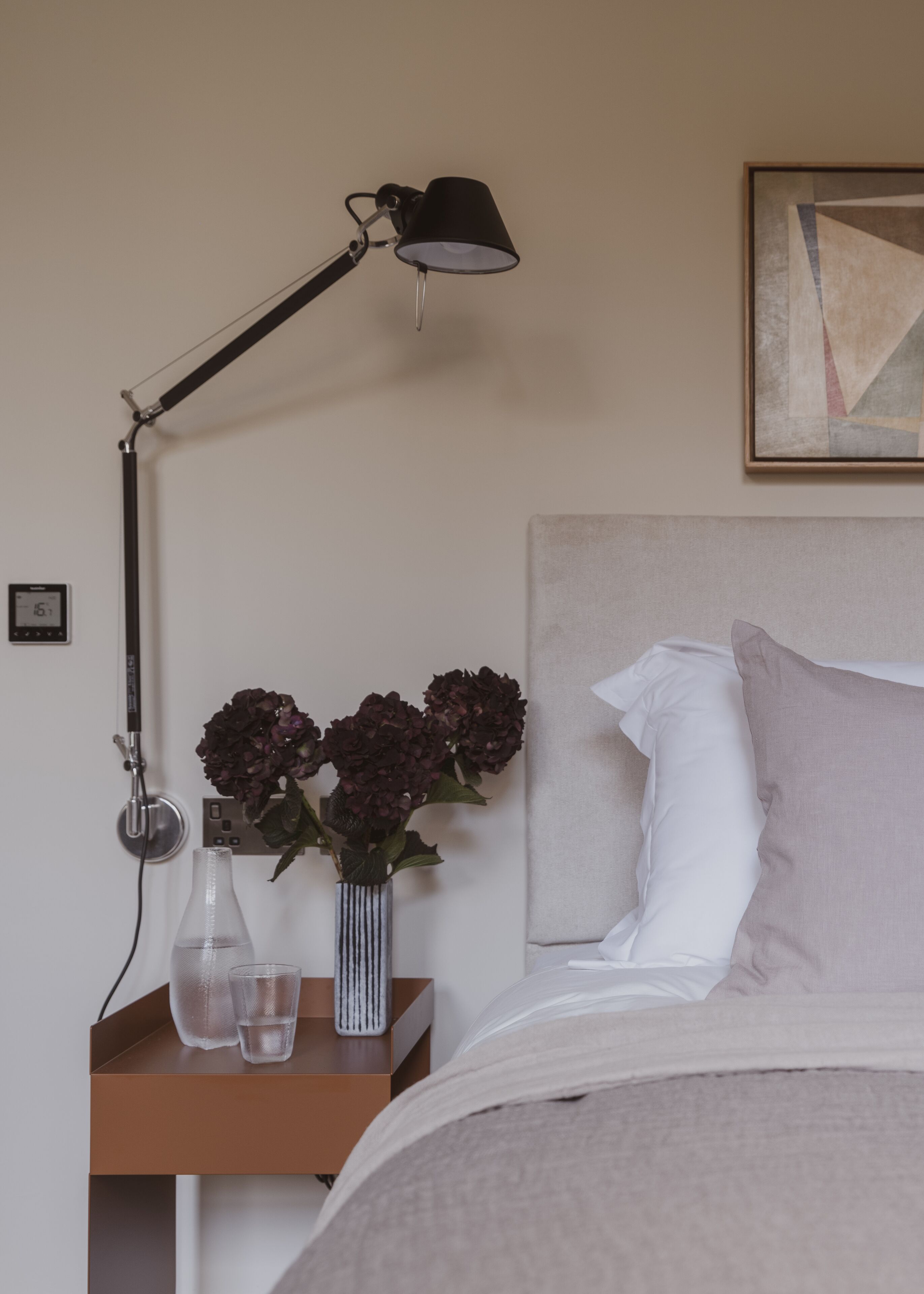 A neatly arranged bedroom with a bed, a bedside table with a lamp, a vase with flowers, and a wall clock.