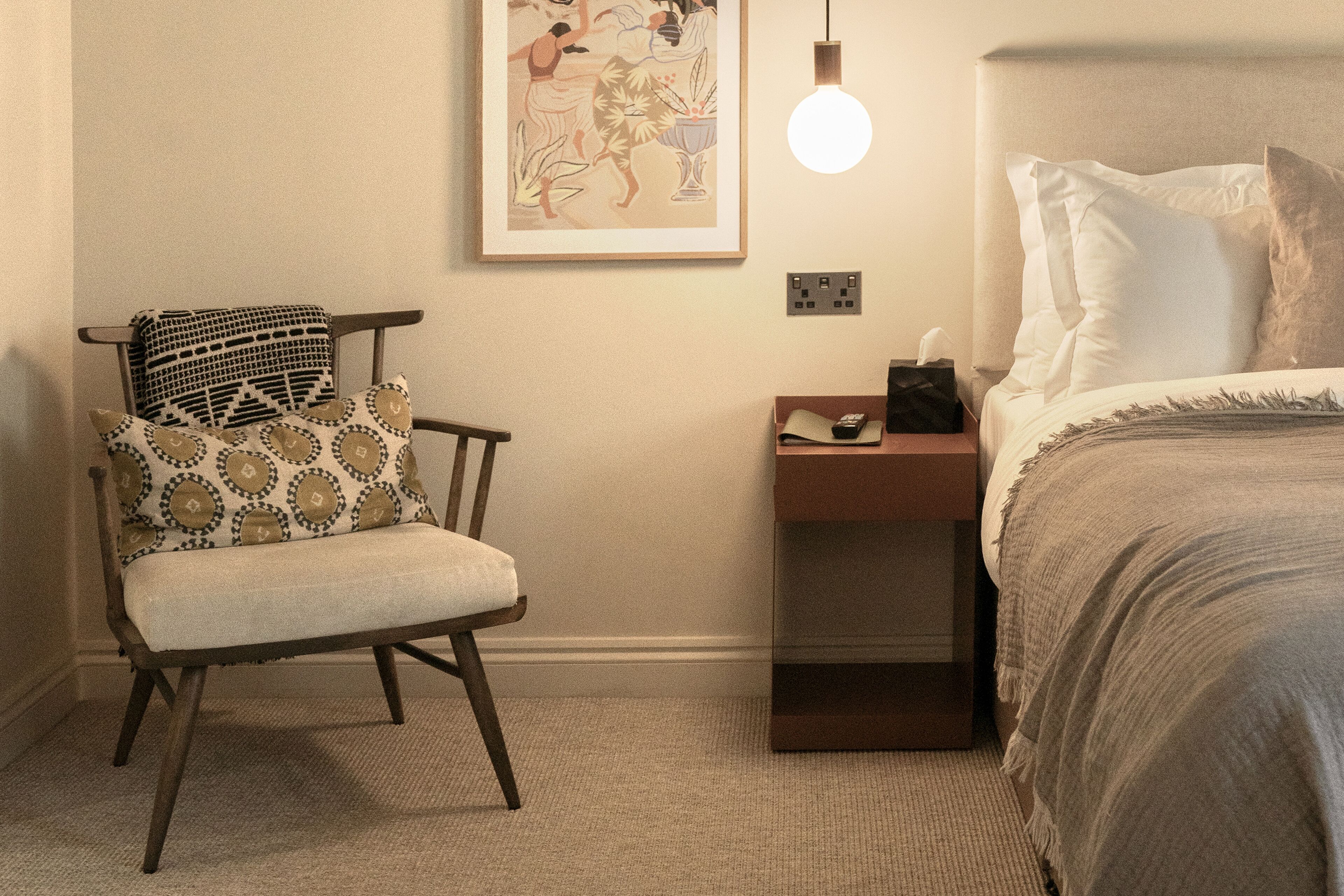 Een slaapkamer met een bed, een stoel met gestreepte kussens, een nachtkastje met een lamp en een ingelijst kunstwerk aan de muur.