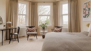 Quarto de hotel com uma cama grande, duas poltronas, uma mesinha e uma janela com cortinas.
