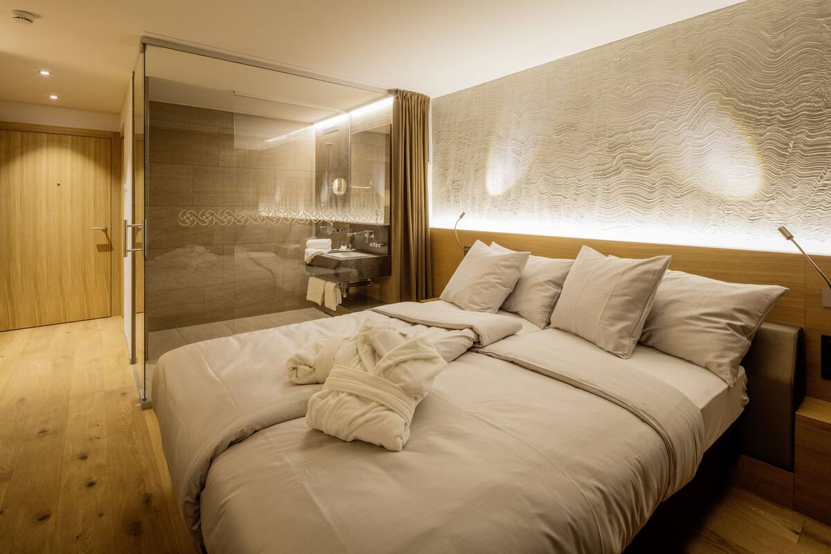 A modern hotel room with a large bed, a walk-in shower, a vanity with a mirror, and a textured wall.