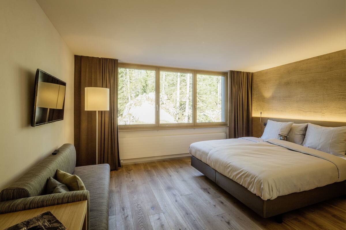 A hotel room with a bed, a sofa, a TV, and a window with curtains.