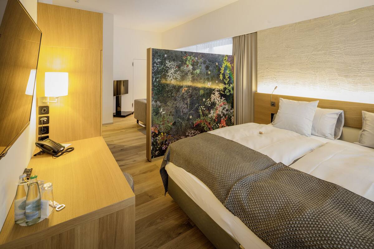 A hotel room with a large bed, a wooden desk with a phone and a lamp, and a view of a forest through a glass partition.
