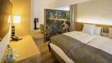 A hotel room with a large bed, a wooden desk with a phone and a lamp, and a view of a forest through a glass partition.