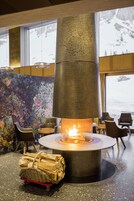 Un interior moderno con una chimenea de piedra central, zona de estar y vistas a montañas nevadas a través de grandes ventanales.