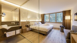 Habitación de hotel moderna con un separador de vidrio, una cama grande, un sofá, un escritorio y una ventana con vista a árboles.