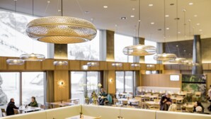 Interior moderno de restaurante con techos altos, lámparas colgantes y una variedad de opciones de asientos, incluyendo cabinas y mesas.