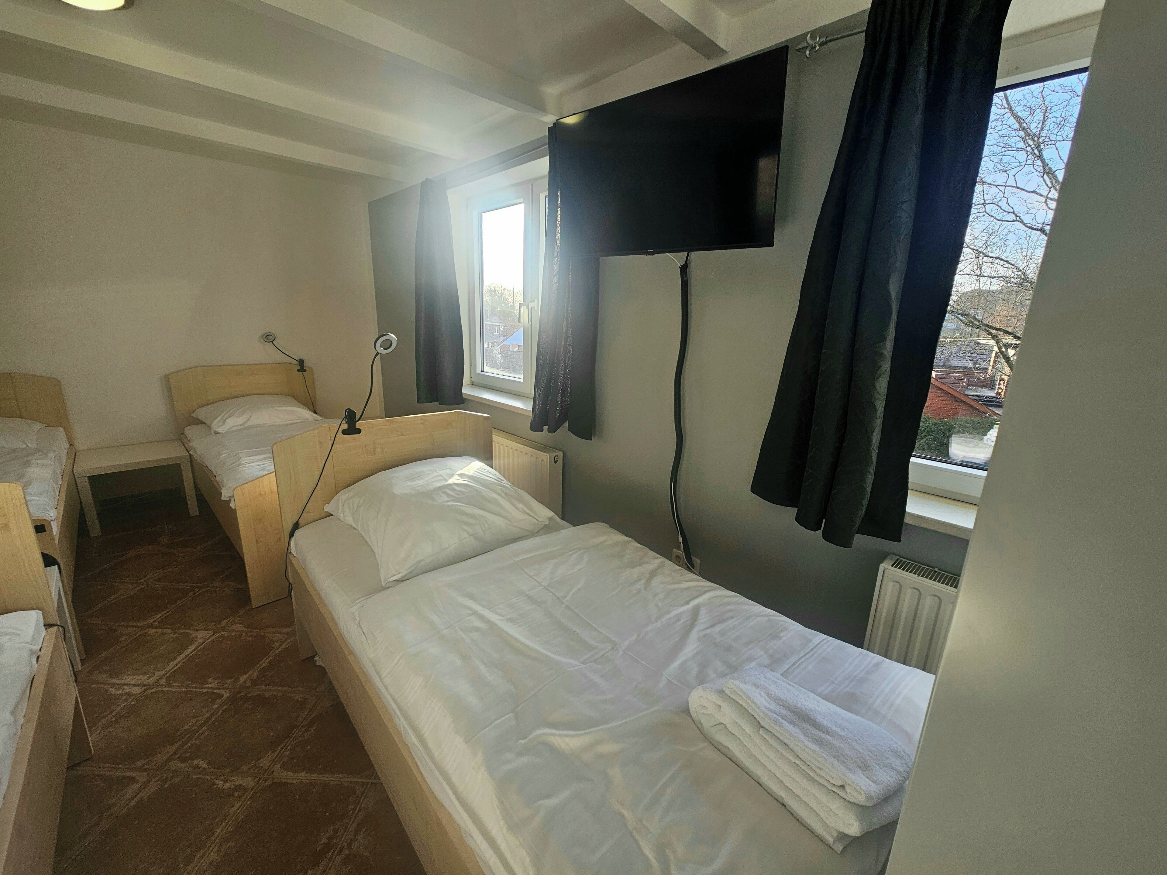 A small, tidy room with a single bed, a bedside table, a window with curtains, and a flat-screen TV mounted on the wall.