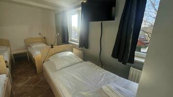 A small, tidy room with a single bed, a bedside table, a window with curtains, and a flat-screen TV mounted on the wall.