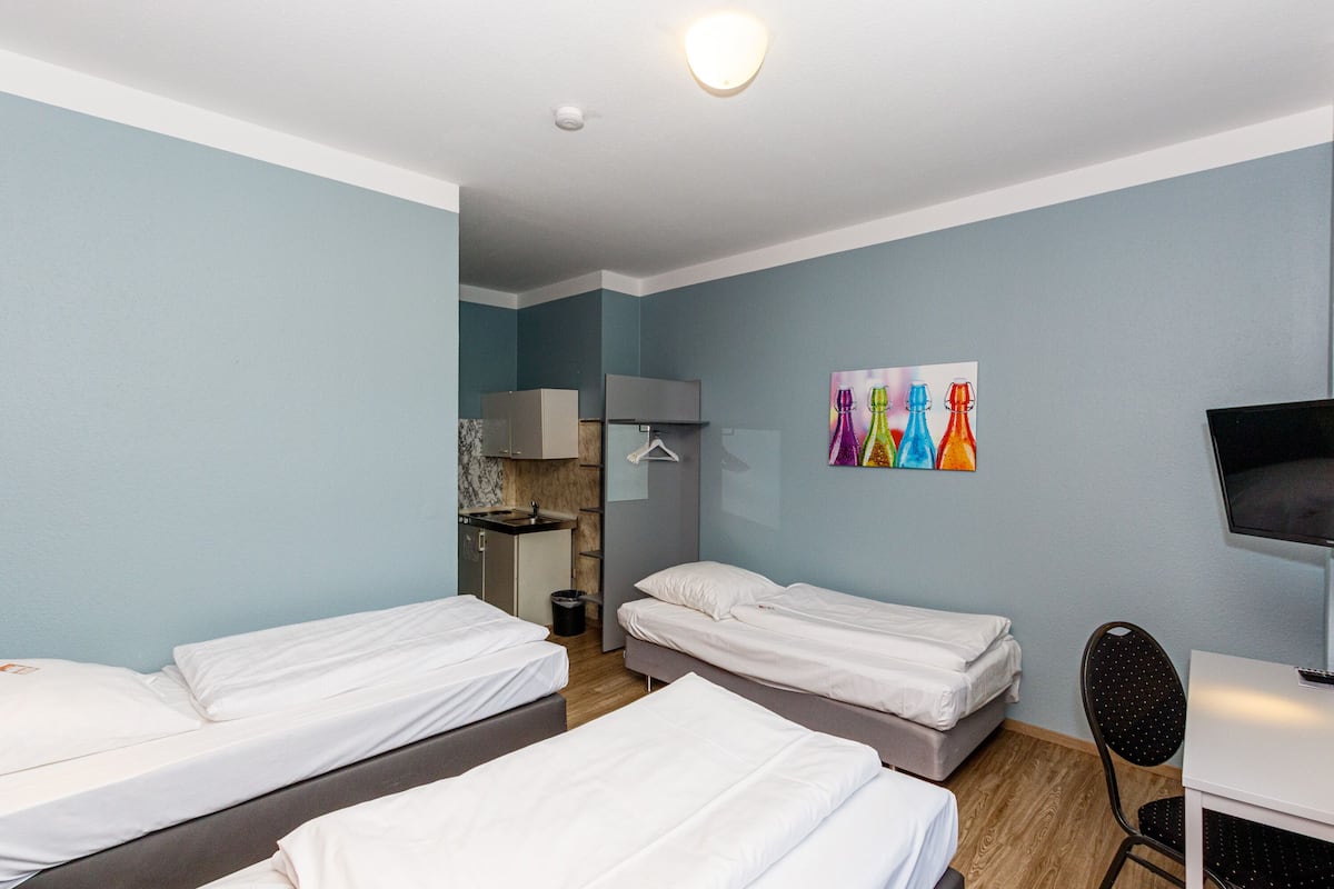 A hotel room with three single beds, a desk with a chair, a television, a small kitchenette, and a colorful artwork on the wall.