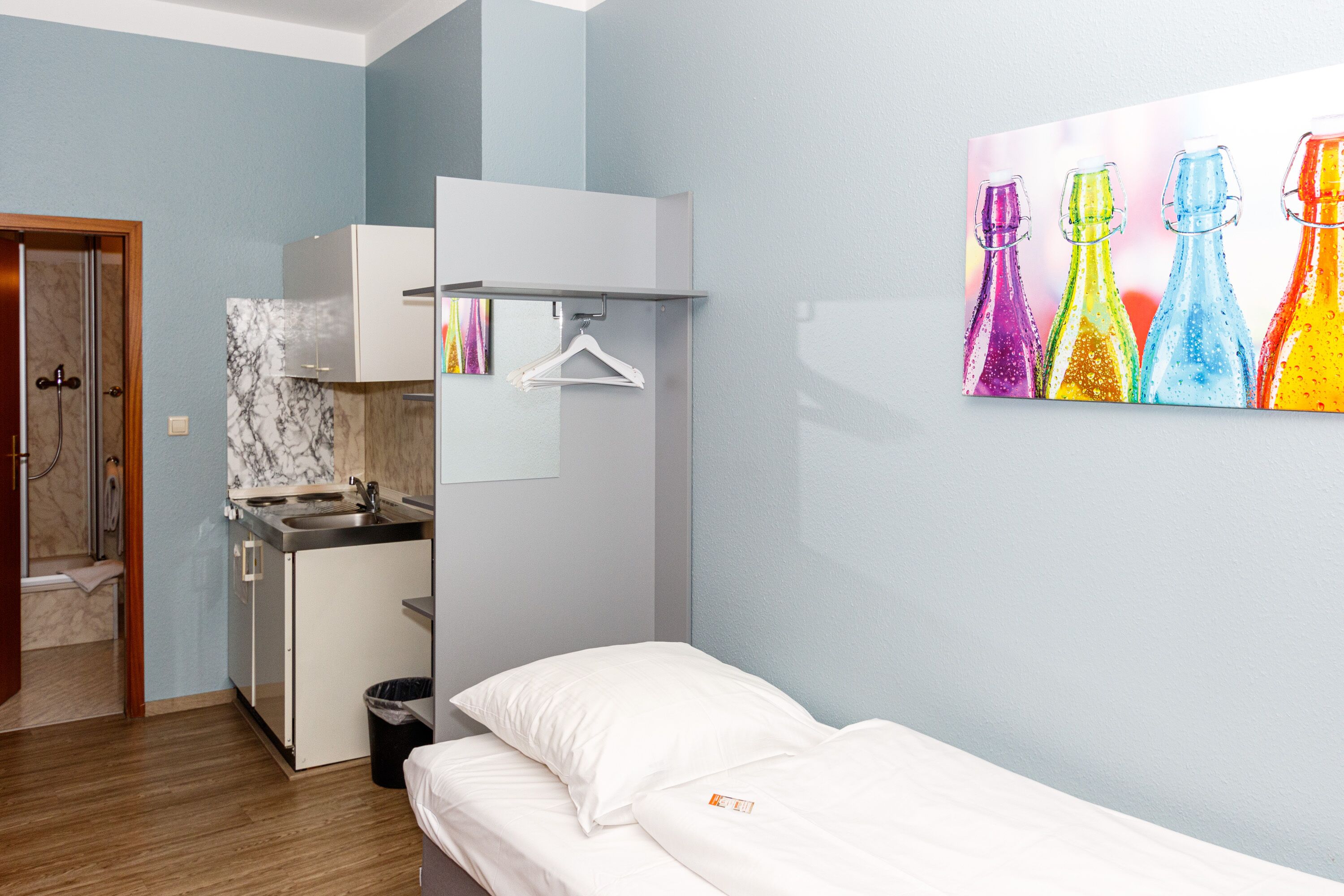 Ein kompaktes Hotelzimmer mit Küchenzeile, einem Bett mit weißer Bettwäsche, einer Wand mit Marmorfliesen und einem farbenfrohen Bild von Flaschen.
